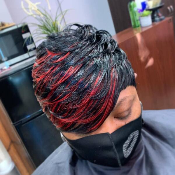 Layered Hairstyles with Streaks of Red Color- a black woman wearing a black face mask and a barber's cape